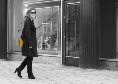 Sonnenbrille tragende Frau auf Stadtbummel mit schwarzen Stiefeln und Mantel vor modernen Ladenfassade in Schwarz-Weiß-Aufnahme.