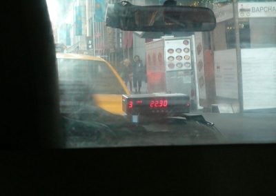 Ruhige Straßenansicht in New York City, Blick durch Taxi-Windschutzscheibe, Stadtmitte mit Passanten und Essenständen, urbanes Straßenbild mit digitaler Uhranzeige, typische Szenerie in Manhattan.