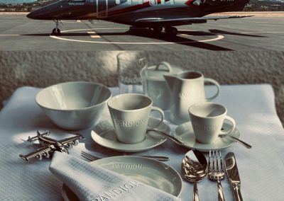 Luxusflugzeug auf der Startbahn mit Blick auf Berge, hochwertiges Geschirr-Set mit Kaffeetassen, Milchkanne, Schüssel, Besteck und Serviette auf einem eleganten Frühstückstisch, Markenlogo "Pandion" sichtbar.
